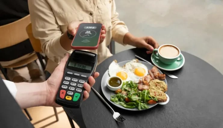A restaurant guest paying at their table with a contactless tap-to-pay phone while a server holds a handheld card reader, beside a brunch plate and a cappuccino.